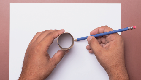 Imagem: Fotografia. Destaque de duas mãos. Uma segura um rolo de papel higiênico vazio e a outra um lápis. Estão sobre uma folha sulfite. Fim da imagem.