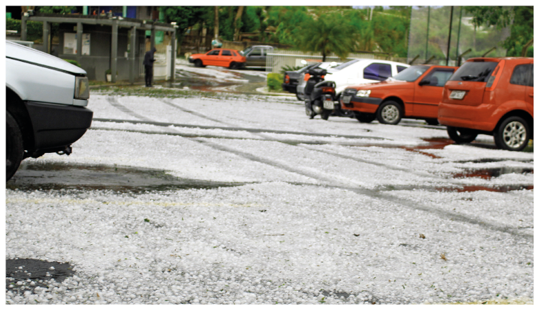 Imagem: Fotografia. Vista de estacionamento de carros com granizo no chão.  Fim da imagem.