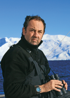 Imagem: Fotografia. Homem de cabelo curto castanho, vestindo casaco preto. Está segurando um binóculo. Atrás há mar e geleiras. Fim da imagem.