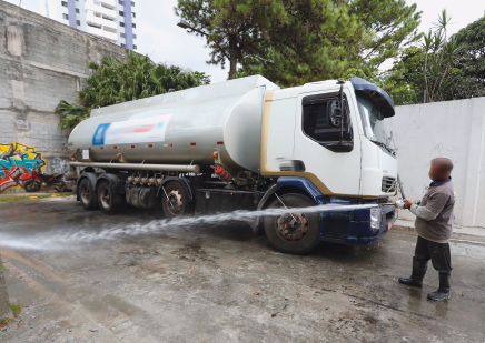 Imagem: Fotografia. Homem calvo, vestindo camiseta cinza e calça preta. Está segurando uma mangueira ligada a um caminhão pipa que transporta água. Está despejando água sobre uma calçada. Fim da imagem.