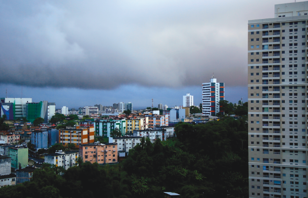 Imagem: Fotografia. Vista de cidade com aglomerado de prédios. Acima, formação de nuvens grandes escuras de chuva. Fim da imagem.