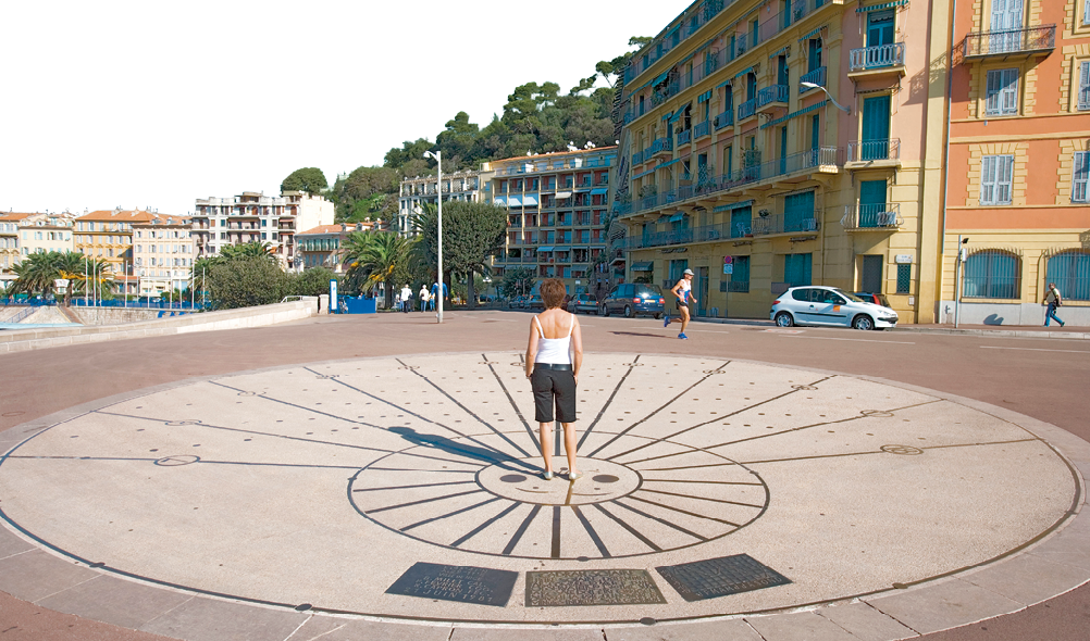 Imagem: Fotografia. Destaque de relógio de sol sobre o chão com marcações. No centro está uma mulher de cabelo curto, vestindo camiseta branca e bermuda preta, está de costas para fotografia. Ao redor da praça há prédios e algumas árvores. Fim da imagem.