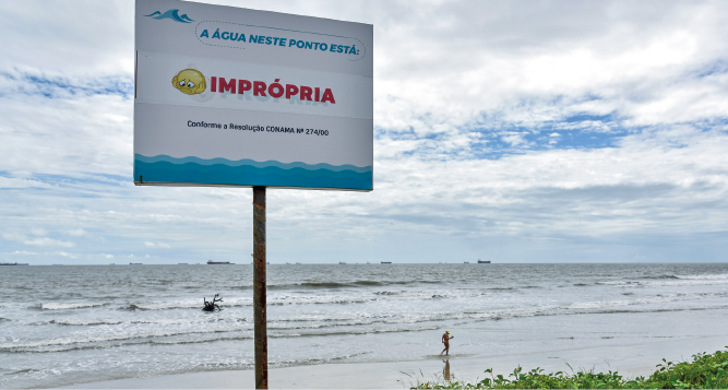 Imagem: Fotografia. Vista de praia com placa em destaque dizendo “a água neste ponto está imprópria”. Fim da imagem.