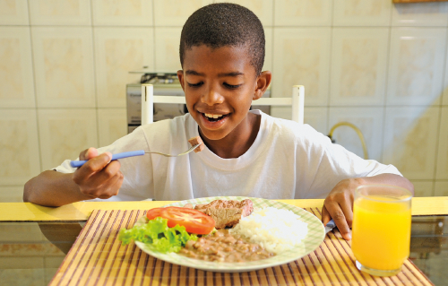 Imagem: Fotografia. Menino de cabelo curto preto, vestindo camiseta branca. Está comendo comida em um prato. Fim da imagem.