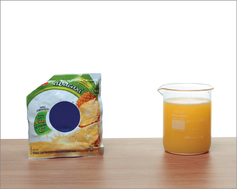 Imagem: Fotografia. Saco de suco em pó. Ao lado, copo com suco laranja. Fim da imagem.