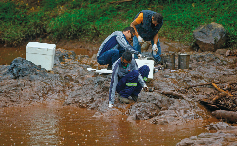 Imagem: Fotografia. Homens sobre a margem de um rio com instrumentos de coleta e transporte de amostras de água. Fim da imagem.