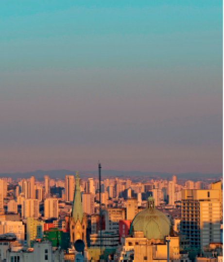 Imagem: Fotografia. Vista de céu em tons de azul e roxo com telhados de prédios iluminados pelo sol. Fim da imagem.