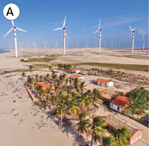 Imagem: Fotografia. A: Vista de vilarejo ao lado de campo desértico com usinas de energia eólica formada por gigantes cata-ventos. Fim da imagem.
