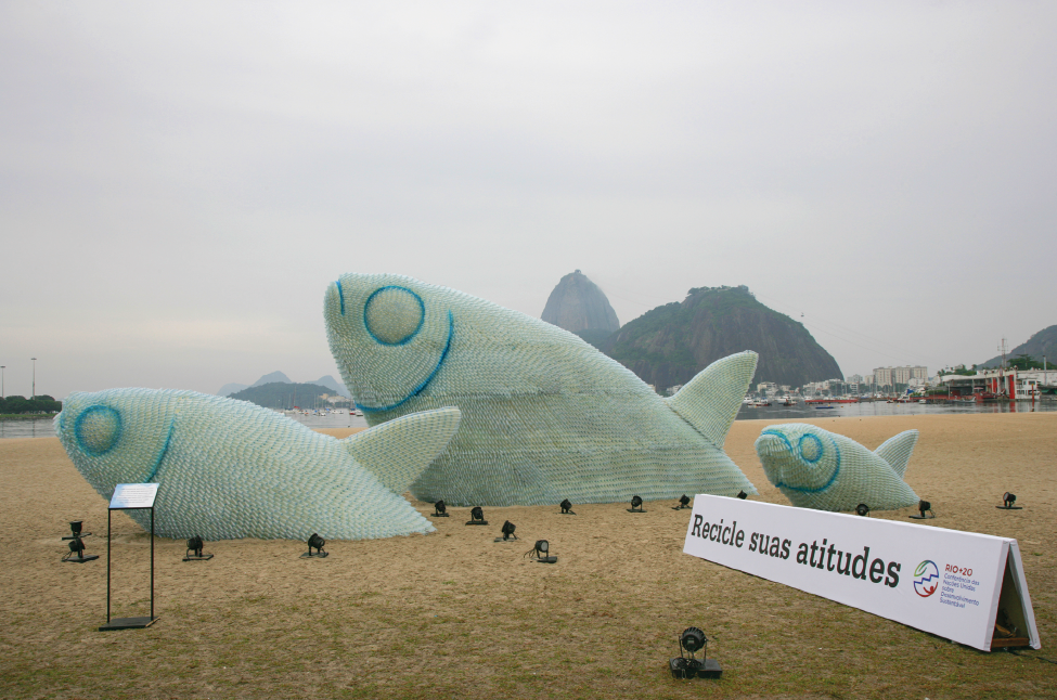 Imagem: Fotografia complementar das páginas 40 e 41. Praia com duas esculturas gigantes de três peixes com holofotes nos arredores. Ao lado, título “recicle suas atitudes”. Fim da imagem.