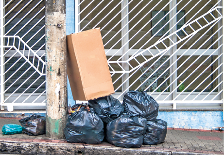Imagem: Fotografia. Lixo domiciliar. Vista de sacos de lixo e caixas de papelão em frente a uma casa. Fim da imagem.