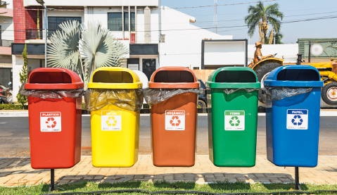 Imagem: Fotografia. Vista de cinco latas de lixo coloridas com símbolos de reciclagem. Fim da imagem.