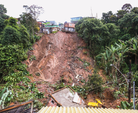 Imagem: Fotografia. Vista de desabamento de morro derrubando casas e destruindo vegetação. Fim da imagem.