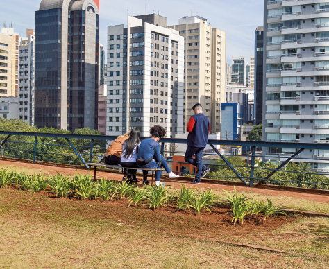 Imagem: Fotografia. Pessoas sentadas em banco de um jardim suspenso com grade de contenção. Ao redor há prédios.  Fim da imagem.