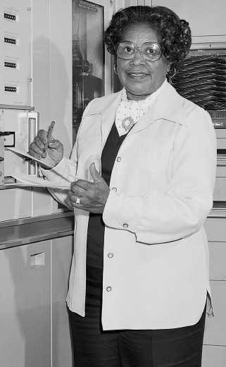 Imagem: Fotografia em preto e branco. Mulher de cabelo curto cacheado e óculos de armação arredondada, vestindo vestido e jaleco. Está segurando uma caneta e uma prancheta. Fim da imagem.