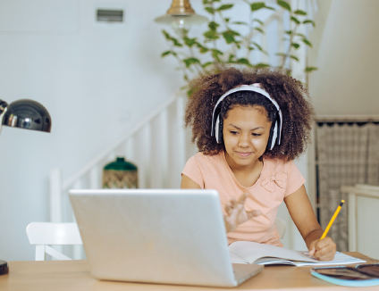 Imagem: Fotografia. Menina de cabelo cacheado longo castanho e fone tampando as orelhas, está segurando um lápis em cadernos. À frente, um computador aberto. Fim da imagem.