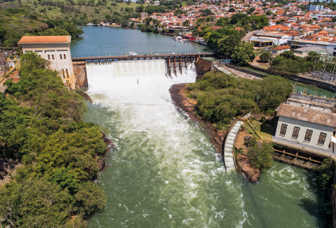 Imagem: Fotografia. Barragem de rio largo com cidade nas margens do rio. Fim da imagem.