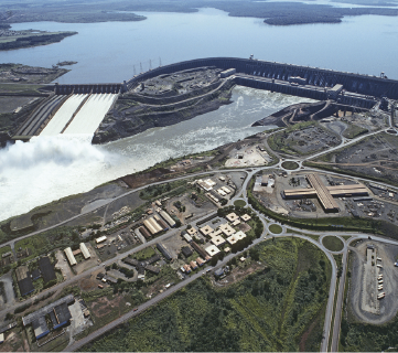 Imagem: Fotografia. Vista aérea de represa em barragem de hidrelétrica. Ao lado há uma cidade Às margens. Fim da imagem.