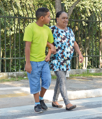 Imagem: Fotografia. Menino de cabelo curto preto, vestindo camiseta verde e bermuda azul. Está de braços entrelaçados com mulher idosa de cabelo longo preto, vestindo camiseta azul e calça cinza. Fim da imagem.