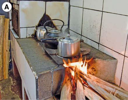 Imagem: Fotografia. A: Fogão a lenha com panelas na parte superior. Na parte inferior há troncos de madeira pegando fogo. Fim da imagem.