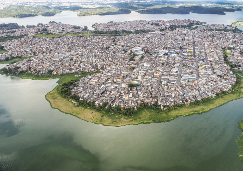 Imagem: Fotografia. Vista aérea de represa com ilha com casas e prédios contemplando todo o território. Fim da imagem.