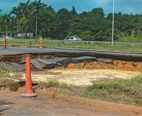 Imagem: Fotografia. Deslizamento formando um buraco sobre pista asfaltada. Há fitas e cones laranjas isolando a área. Fim da imagem.