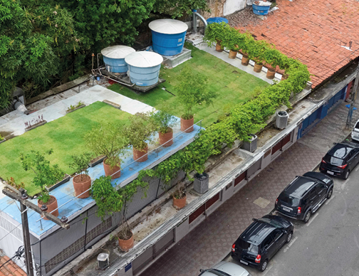 Imagem: Fotografia. Vista aérea de prédio com jardim formado por plantas em vasos e gramado sobre o telhado, ao lado há caixas de água. Sobre a rua há carros estacionados lado. Fim da imagem.