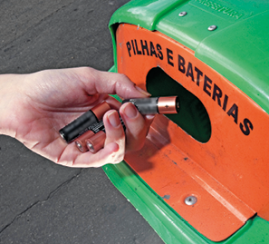 Imagem: Fotografia. Lixeira de descarte indicando “pilhas e baterias”. Destaque de uma mão jogando pilhas. Fim da imagem.