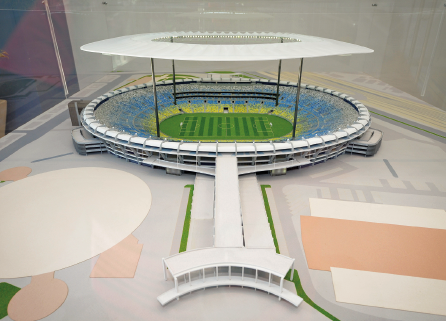 Imagem: Fotografia. Vista de maquete de um estádio de futebol. Passarela longa com ligação da abertura até a arquibancada arredondada. Fim da imagem.