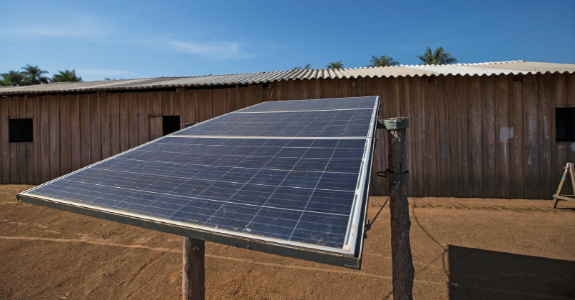 Imagem: Fotografia. Placa de coleta de energia solar ao lado de um barracão. Fim da imagem.