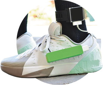 Imagem: Fotografia. Destaque de um pé com tênis branco com um marcador no tornozelo e uma bateria ligada ao tênis. Fim da imagem.