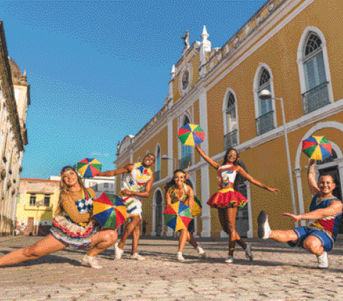 Imagem: Fotografia. Cinco pessoas estão segurando guarda-chuvas pequenos e coloridos e dançando. Atrás delas há uma construção grande com paredes amarelas. Fim da imagem.