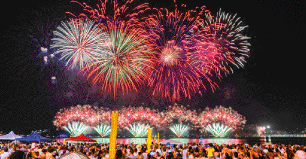 Imagem: Fotografia. Uma multidão aglomerada. Ao fundo, fogos de artifício coloridos no céu escuro. Fim da imagem.