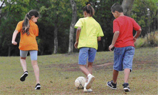 Imagem: Fotografia. Duas meninas e um menino estão de costas e correndo atrás de uma bola de futebol. Ao fundo, árvores. Fim da imagem.