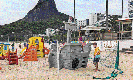 Imagem: Fotografia. Crianças em volta de brinquedos na areia da praia. À direita, um menino está na frente de um brinquedo cinza com formato de navio. Ao fundo, morros e árvores.  Fim da imagem.