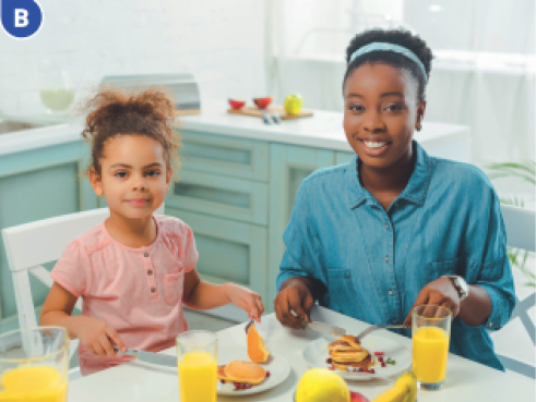 Imagem: Fotografia B. A menina está sentada ao lado de uma mulher. Entre elas há uma mesa com comida em cima.  Fim da imagem.