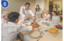 Imagem: Fotografia C. Uma família com roupa branca sentada em volta de uma mesa com comida em cima.  Fim da imagem.