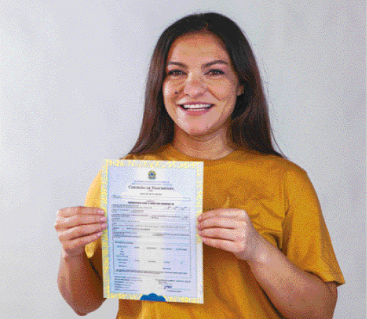 Imagem: Fotografia. Mulher com cabelo liso e comprido e camiseta amarela está sorrindo e segurando uma certidão de nascimento. Fim da imagem.