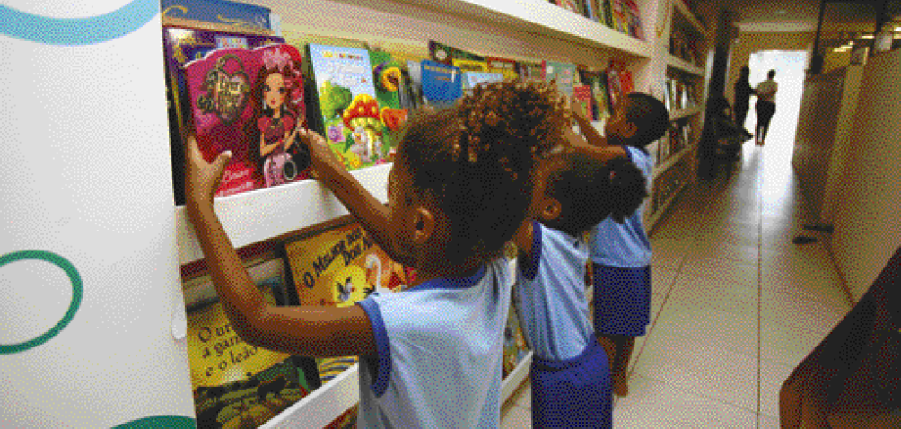 Imagem: Fotografia. Três crianças com camiseta azul estão de costas e segurando livros sobre uma prateleira. Ao fundo, mais prateleiras com livros. Fim da imagem.