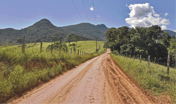 Imagem: Fotografia. No centro, estrada de terra e nas laterais, plantas e árvores. Ao fundo, morros e nuvens brancas no céu azul. Fim da imagem.