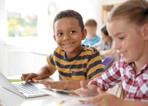 Fotografia. O menino está sentado com as mãos sobre o teclado de um notebook. Ao seu lado, uma menina está sentada e sorrindo.