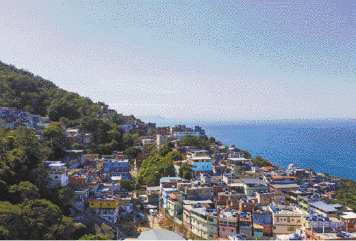 Imagem: Fotografia. Vista aérea de casas e árvores na encosta de um morro. Ao lado, o mar. Fim da imagem.