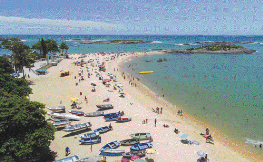 Imagem: Fotografia. Vista aérea de barcos e pessoas na areia da praia. Ao lado há várias árvores. Ao fundo, o céu claro.  Fim da imagem.