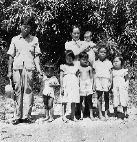 Imagem: Fotografia em preto e branco. Um casal e cinco crianças estão em pé e olhando para frente. A mulher está segurando um bebê no colo e ao fundo há árvores.  Fim da imagem.