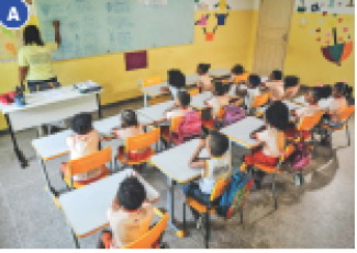 Imagem: Fotografia A. Crianças sentadas em carteiras escolares. Na frente delas, uma professora está escrevendo na lousa.  Fim da imagem.