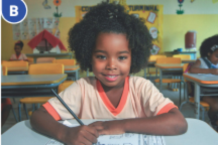 Imagem: Fotografia B. Menina com camiseta laranja está sentada em uma carteira escolar e segurando um lápis sobre um papel.  Fim da imagem.