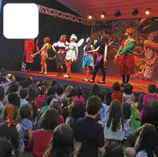 Imagem: Fotografia. Pessoas sentadas em poltronas. Ao fundo, pessoas fantasiadas sobre um palco. Fim da imagem.