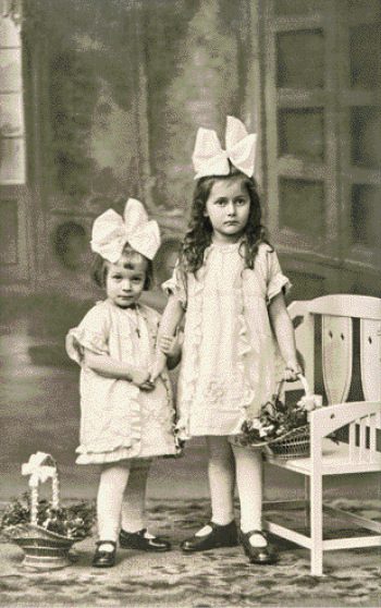 Imagem: Fotografia em preto e branco. Duas meninas com laço grande na cabeça e vestido estão em pé e olhando para frente.  Fim da imagem.