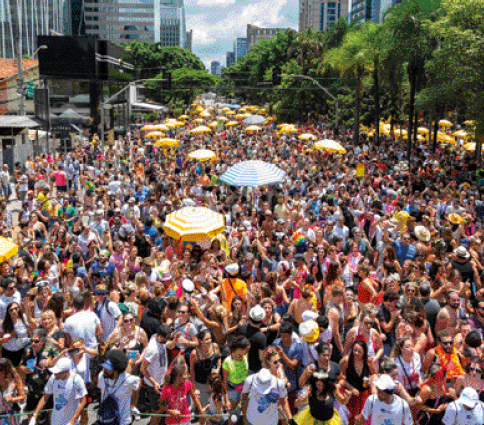 Imagem: Fotografia. Uma multidão aglomerada em uma rua. Nas laterais há árvores e ao fundo, prédios. Fim da imagem.