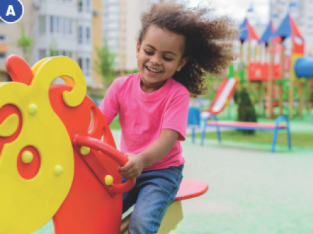 Imagem: Fotografia A. Uma menina com cabelo encaracolado e comprido está sorrindo e sentada em um brinquedo com formato de cavalo. Ao fundo, um escorregador com formato de castelo.  Fim da imagem.