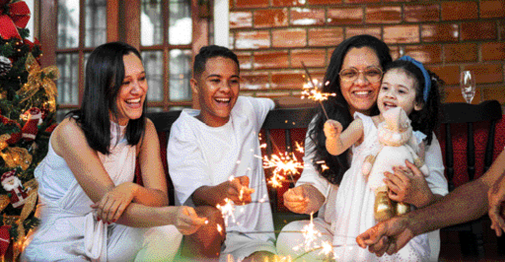 Imagem: Fotografia. Duas mulheres, um menino e uma menina com roupas brancas estão segurando velas de faísca e sorrindo. Ao lado há uma árvore de Natal enfeitada. Fim da imagem.